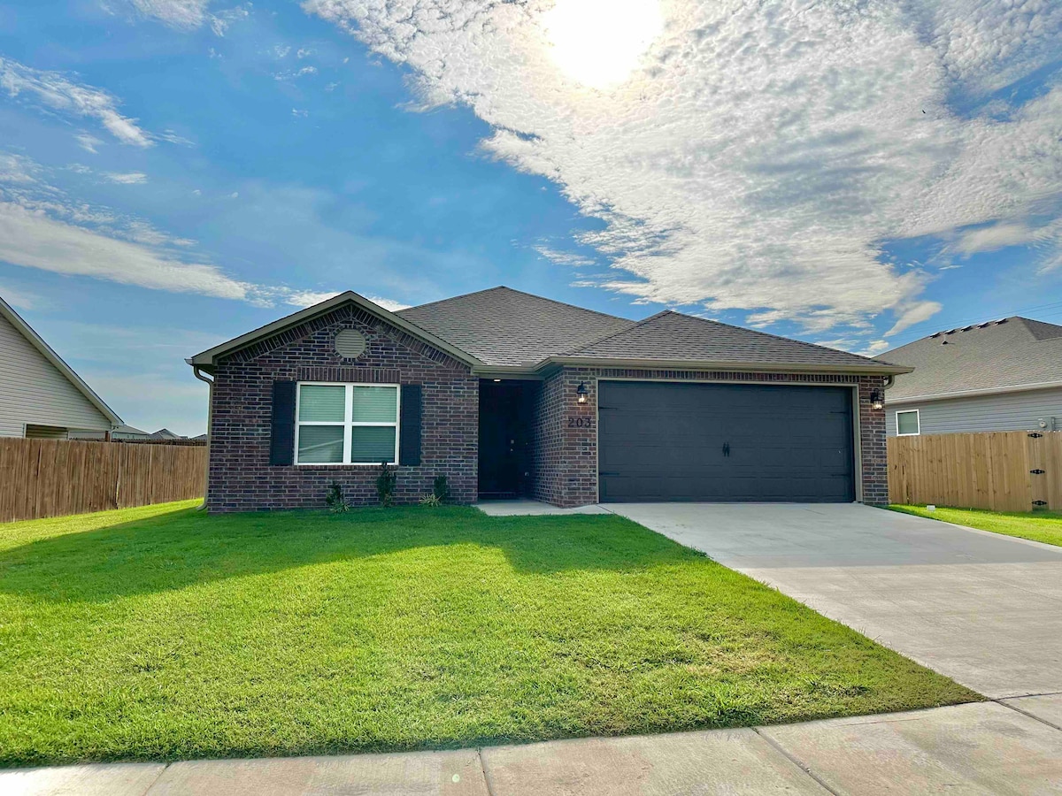 A single-story brick home features a covered entrance and a two-car garage. The well-maintained lawn is bordered by wooden fencing, and the sky is bright with scattered clouds, creating a welcoming exterior view.