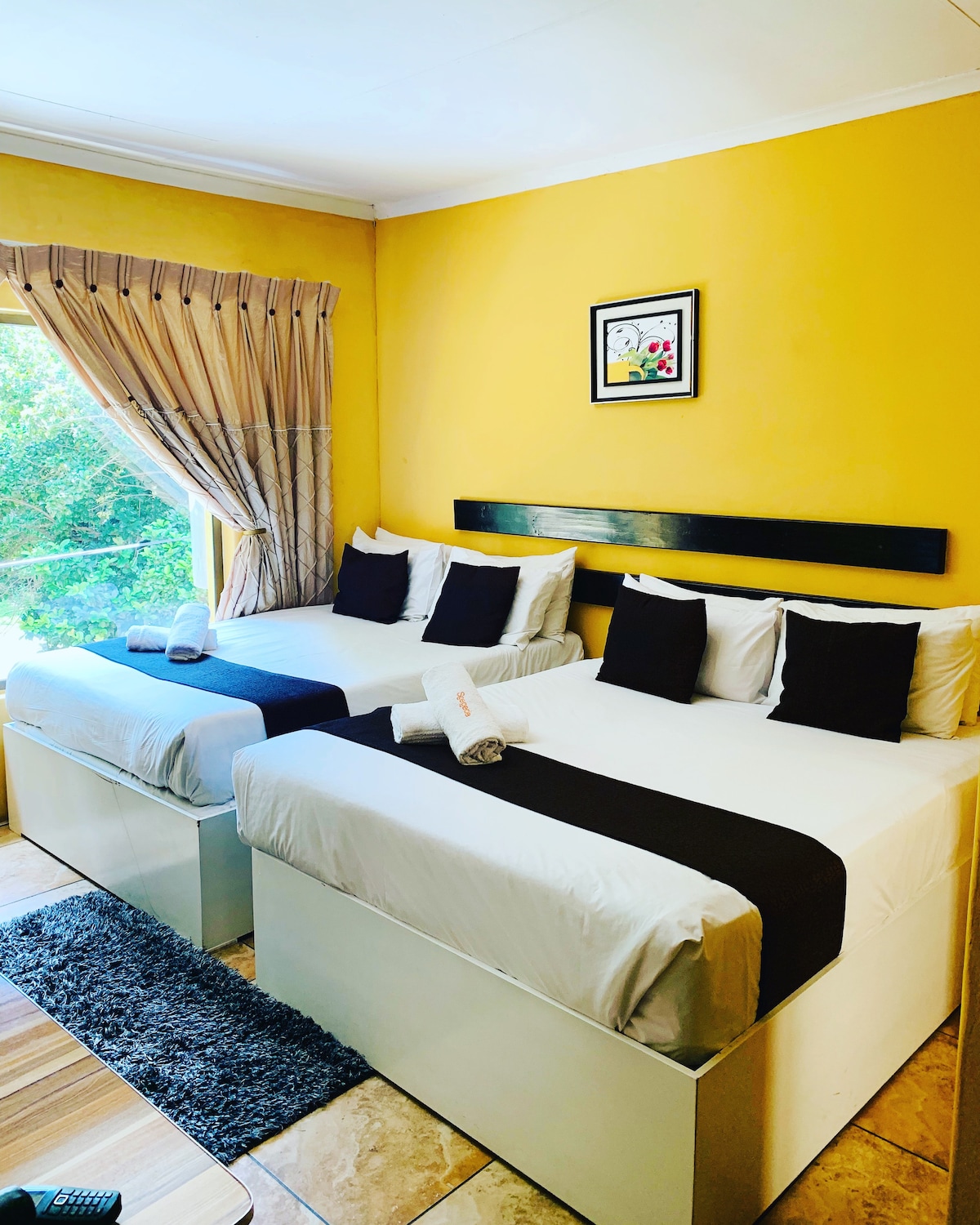 A bright room features two spacious beds dressed in white linens with black accents. A decorative wall piece hangs above the beds. Natural light enters through a large window, complemented by drapes, while light-colored flooring and a patterned rug add warmth to the space.