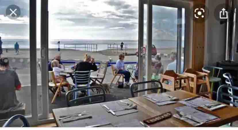 A spacious dining area is visible through large glass doors, showcasing outdoor seating along the beach. Tables are set with utensils, and guests can be seen enjoying their meals while overlooking the water. The relaxed atmosphere is enhanced by natural light filtering through the windows.