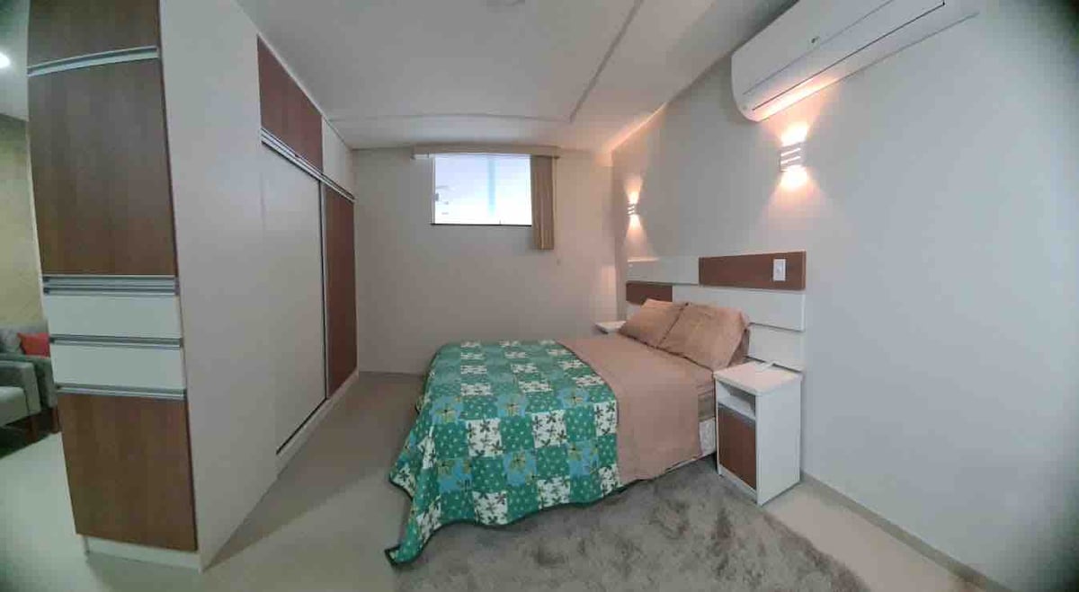 A cozy sleeping area features a spacious bed adorned with a patterned blanket. Nightstands are positioned on either side, and soft lighting is provided by wall sconces. A window allows natural light to filter in, enhancing the neutral tones of the decor.