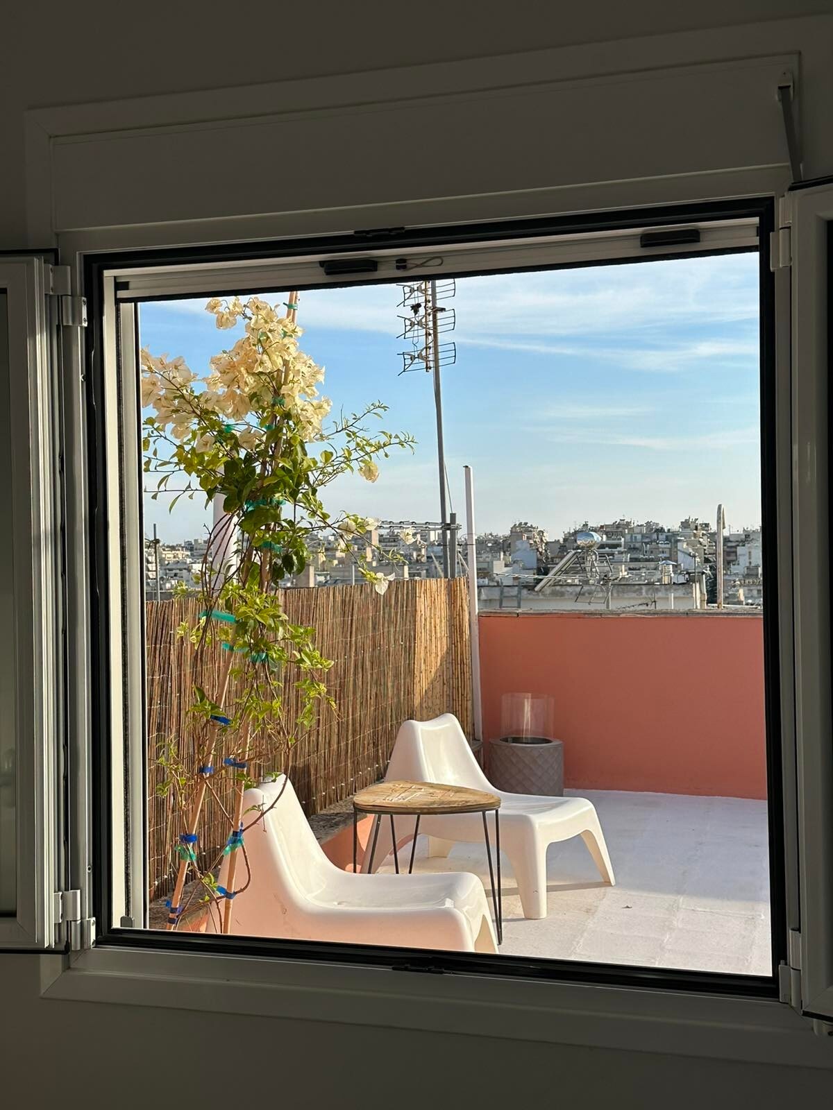 The view from the window opens to a balcony area featuring two white chairs and a small round table. Bougainvillea climbs along the window frame, adding greenery against the backdrop of city buildings and a clear sky.