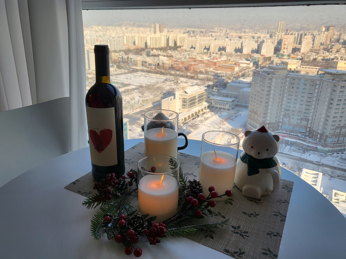 A dining table is set with a bottle of red wine and three candles, each lit softly. Seasonal decor including a small white bear figurine and natural elements can be seen, with a panoramic view of the cityscape visible through the window.