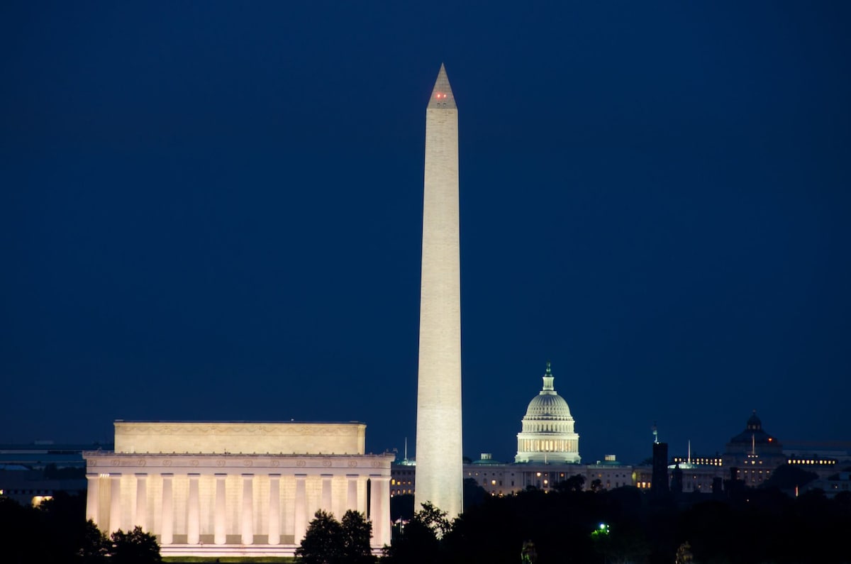 An evening view of iconic DC monuments is presented, highlighting the illuminated Lincoln Memorial in the foreground and the Washington Monument rising prominently. The dome of the U.S. Capitol can be seen in the distance, surrounded by a softly lit night sky.