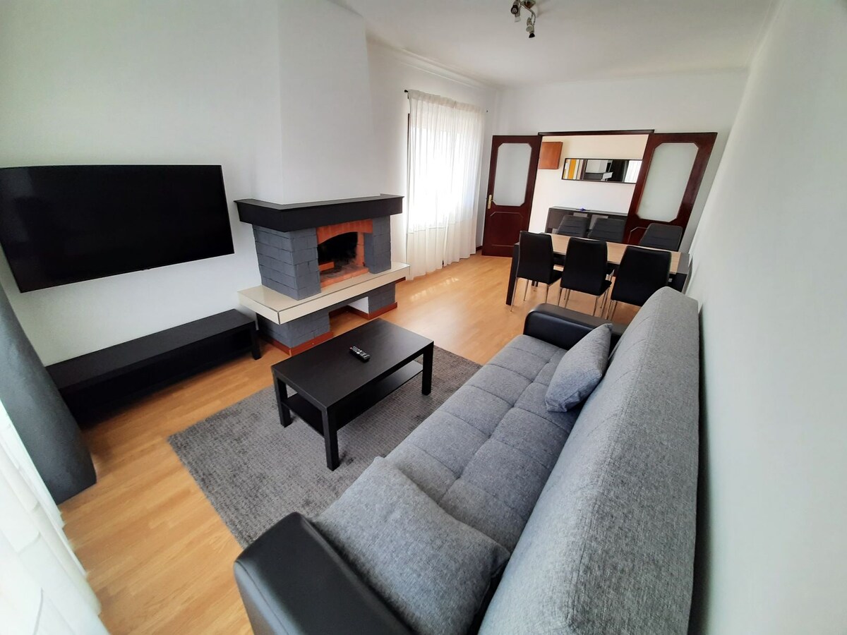 A spacious living area is shown with a grey sectional sofa facing a mounted flat-screen TV. A sleek black coffee table sits on a soft area rug, while a modern fireplace adds warmth. Six dining chairs are arranged around a table, visible in the background.