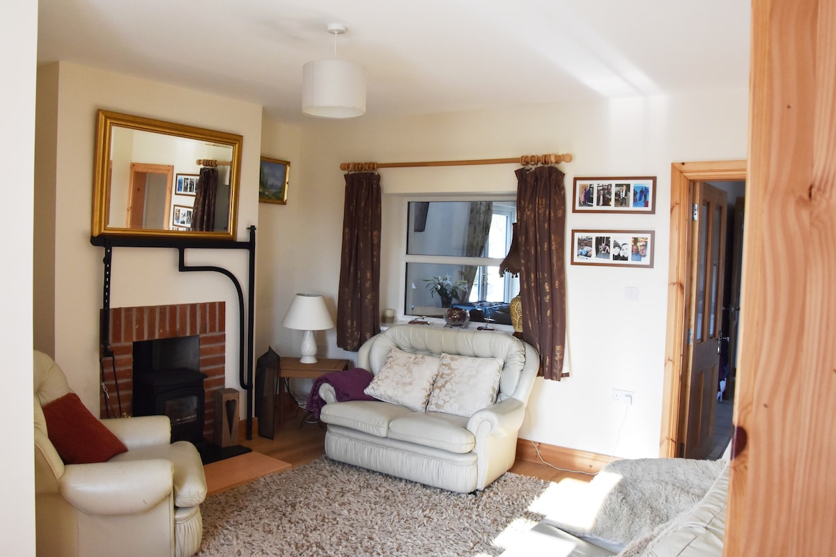 A comfortable lounge area is presented, featuring light-colored leather sofas arranged around a central rug. A brick fireplace is visible, accompanied by a white lamp on a side table. Natural light enters through a window adorned with soft drapes, complemented by framed photos on the wall.