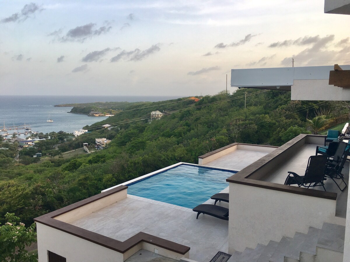 A stunning view features a tranquil infinity pool overlooking the lush green hillside and the bay. Comfortable lounge chairs line the poolside, inviting relaxation. The sky is lightly clouded, reflecting soft colors as the sun sets beyond the horizon.