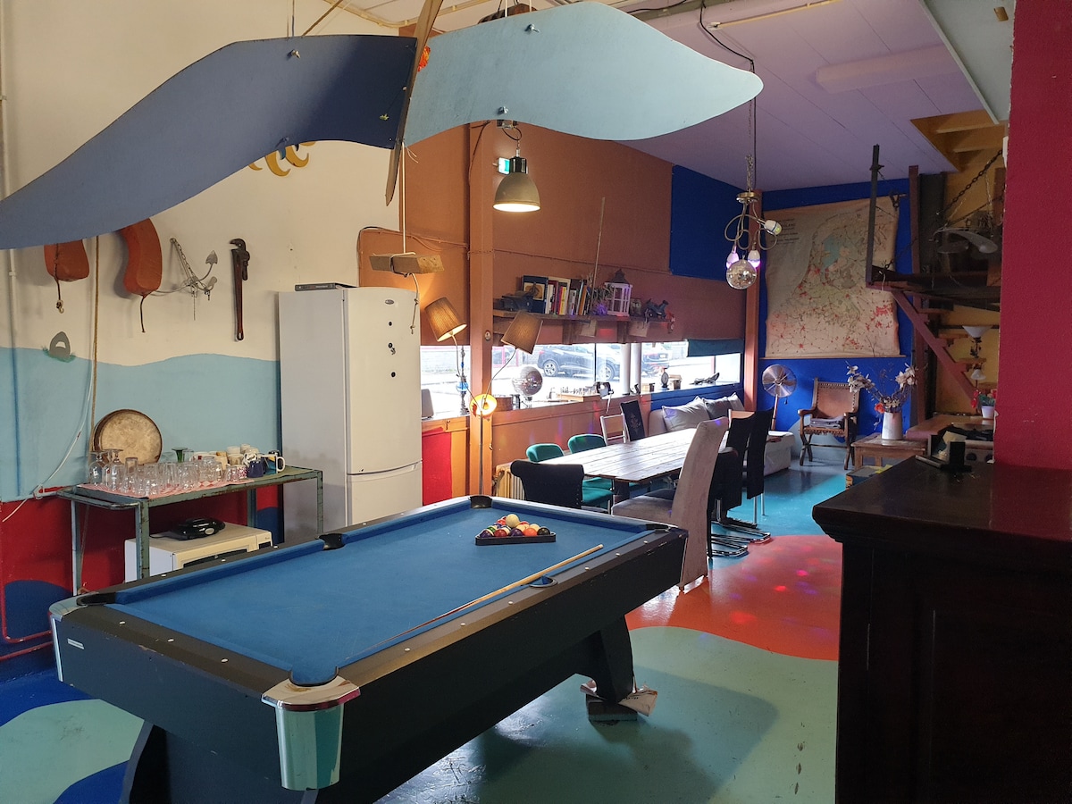 A spacious area is designed with vibrant colors and distinct sections. A billiard table occupies the center, surrounded by various seating options, including a large dining table. A refrigerator and shelves are visible, blending practical elements with artistic decor.