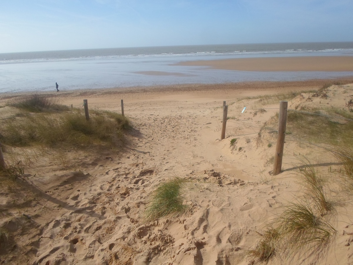 A sandy pathway descends from wooden posts towards a beach, with gentle waves visible in the distance. Dune grasses partially frame the scene, and a lone figure can be seen walking along the shore. The sky is clear, indicating a pleasant weather.