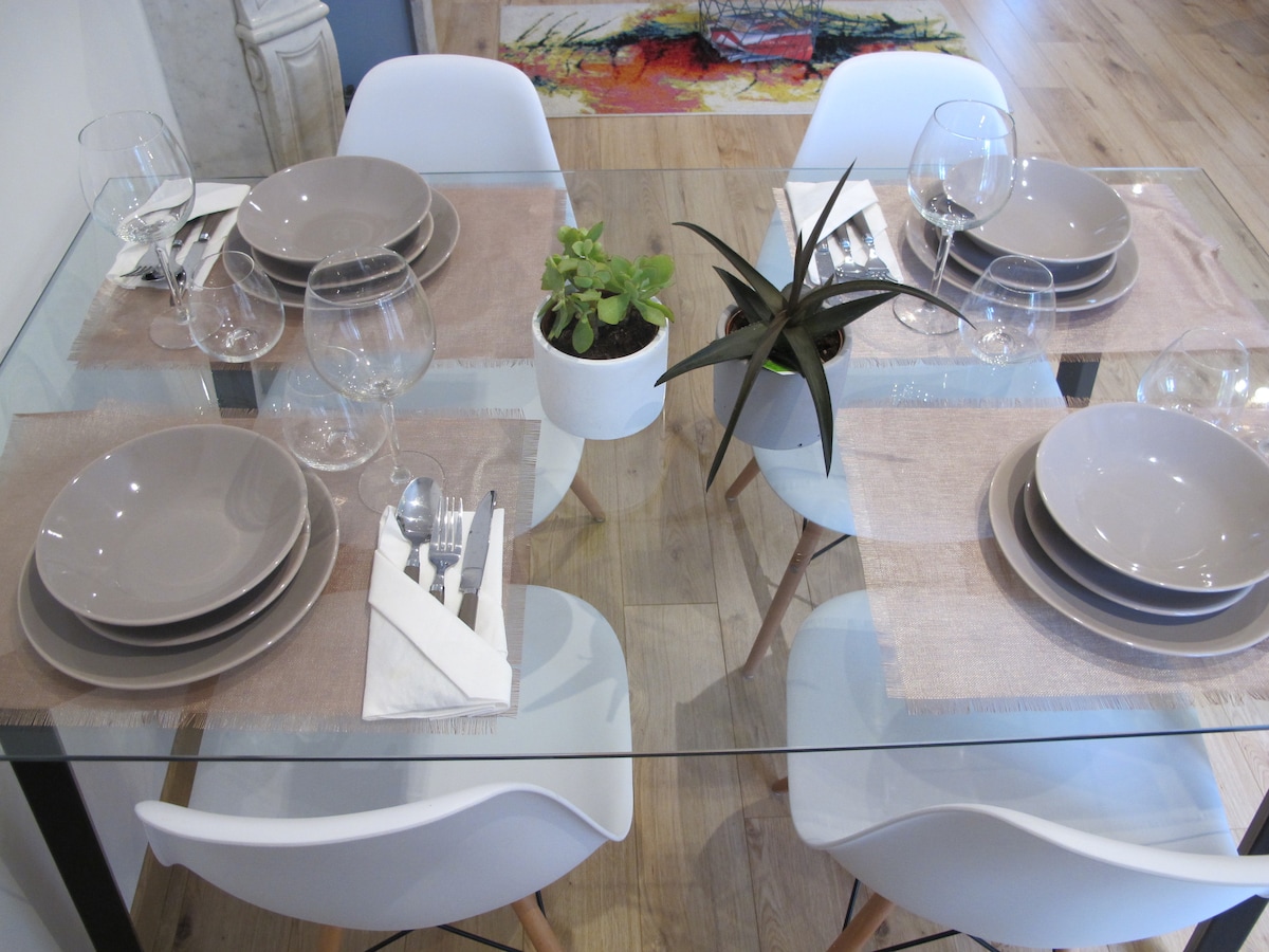 A modern dining table is set for four, featuring glassware, plates, and utensils placed neatly on brown placemats. A small potted plant adds a touch of greenery to the center, while stylish white chairs are positioned around the table.