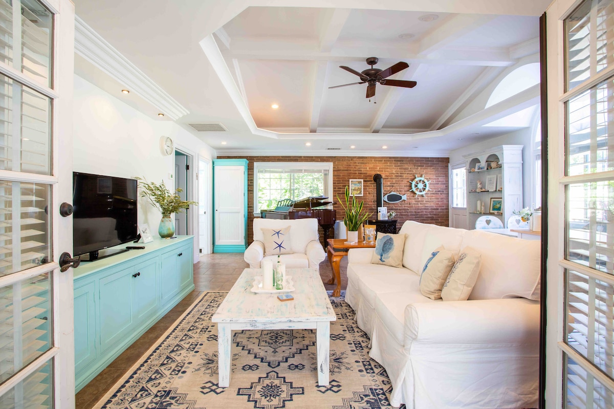 A spacious living area features a comfortable white sofa and a decorative coffee table on a patterned rug. Natural light fills the room through large windows, highlighting a wall-mounted TV and a grand piano in the background.