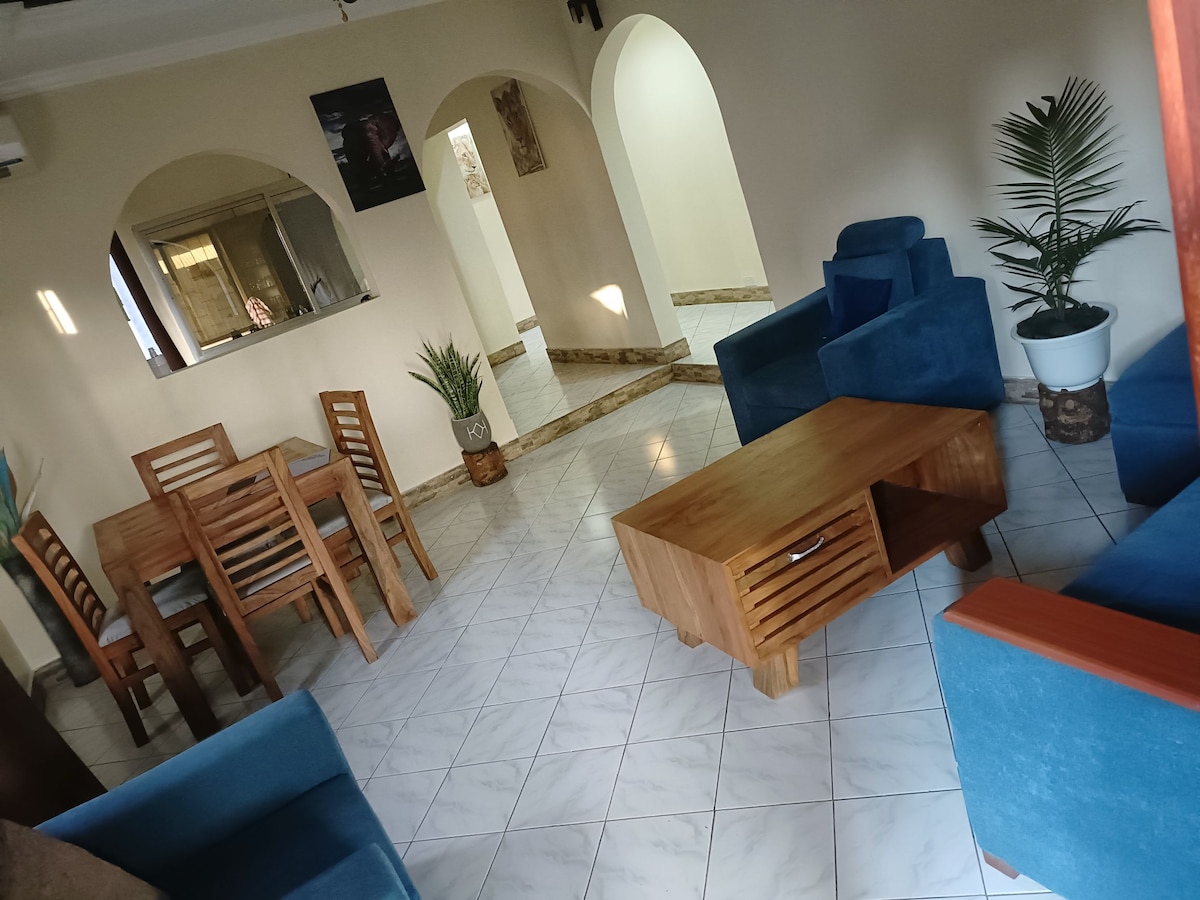 An open living area features blue upholstered seating arrangements paired with a wooden coffee table. A dining set with a wooden table and chairs is visible in the background. Tropical plants are positioned in decorative pots, enhancing the room's natural ambiance.