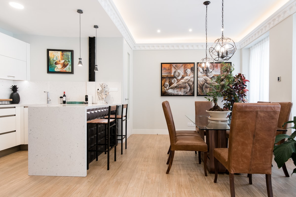 The modern kitchen features sleek white cabinetry and a spacious island with bar seating. A dining area is highlighted by a wooden table surrounded by leather chairs, with artwork on the walls adding a touch of elegance. Natural light enhances the airy atmosphere.