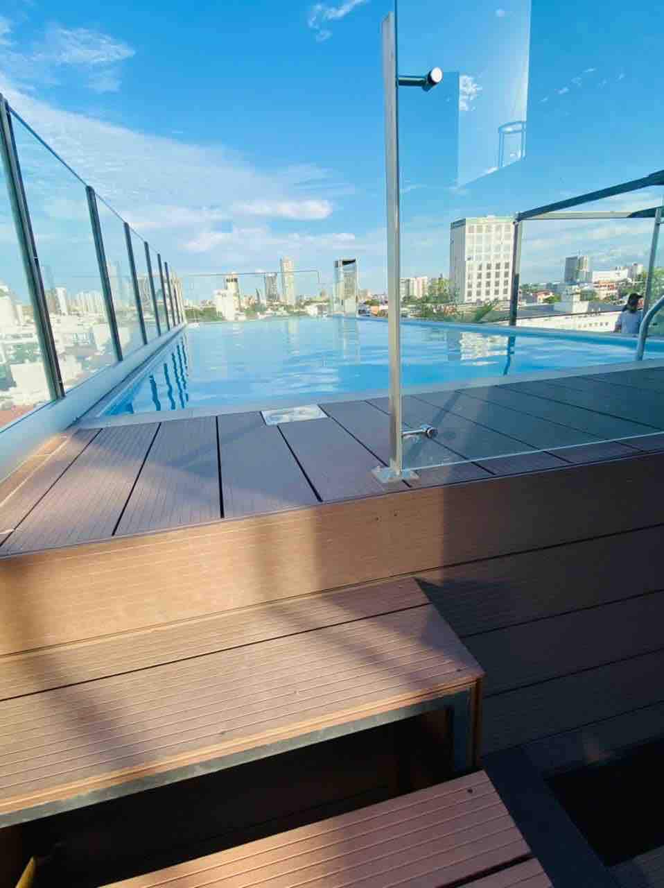 A rooftop pool is visible, bordered by a clear glass railing and wooden decking. Steps lead down into the water, reflecting the sky. A city skyline can be seen in the background under a bright blue sky.