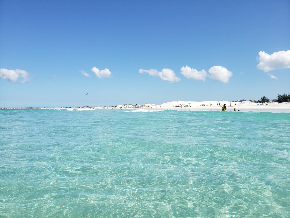 A clear view of the beach features soft white sand dunes under a bright blue sky. The turquoise water is calm, with gentle ripples reflecting the sunlight. Beachgoers are seen enjoying the shoreline in the distance, creating a relaxed atmosphere.
