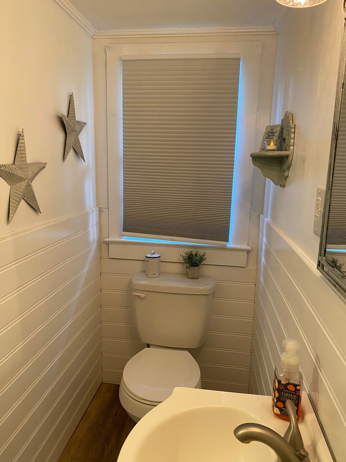 A compact bathroom is presented, featuring a toilet and a small sink. Light filters through a window with a partially closed blind. Decor includes two metal stars on the walls, and a small plant is placed on a shelf above the toilet.