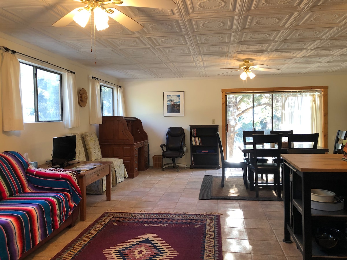 A spacious great room features a mix of seating options, including a striped sofa and a wooden desk. Windows allow natural light to brighten the tiled flooring. A dining table with black chairs occupies one corner, while a cozy reading area is highlighted by a small bookcase.
