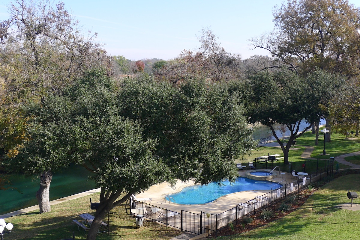 A view of the resort-style pool and hot tub, surrounded by lush greenery. The area is fenced and features lounge chairs positioned around the pool. The backdrop includes a glimpse of the Comal River, enhancing the outdoor experience.