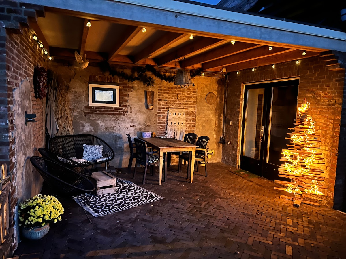An outdoor seating area features a wooden dining table surrounded by black chairs, illuminated by warm string lights. A decorative tree made of wooden planks adds a unique touch, while a patterned rug and potted flowers enhance the ambiance.