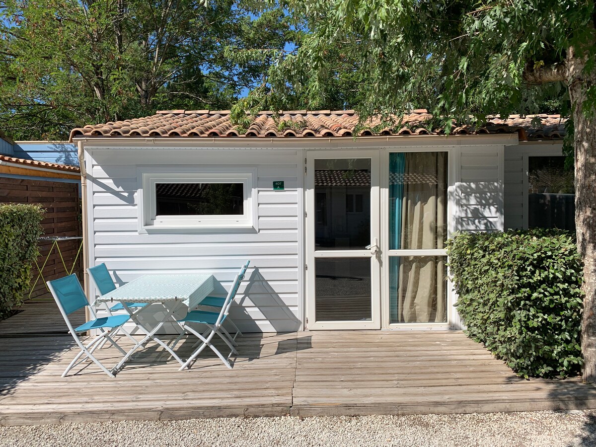 A light-colored chalet exterior is surrounded by greenery, featuring a wooden deck with a table and chairs. Glass doors provide access to the interior, while large windows allow natural light to illuminate the space. The roof showcases traditional tiles.