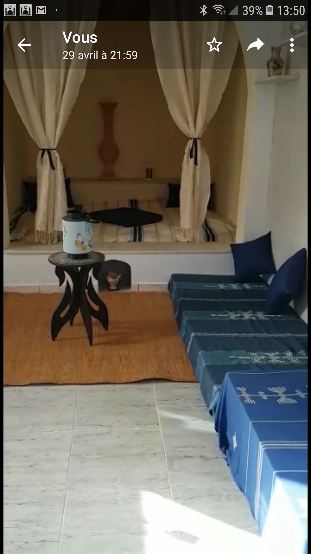 A cozy seating area features plush blue cushions on a textured rug. A small table stands nearby, with a decorative vase displayed on top. Soft drapes frame a serene alcove with a low bed, creating a calming and inviting atmosphere.
