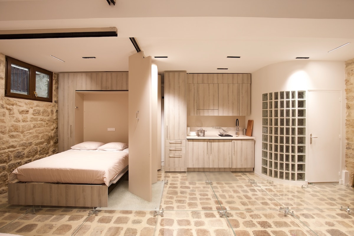 A spacious loft features exposed stone walls and a clear glass floor, revealing 16th-century cobblestones below. A Queen-size bed is integrated within the cabinetry, which also includes a modern kitchen setup. Natural light is provided through windows and architectural accents.