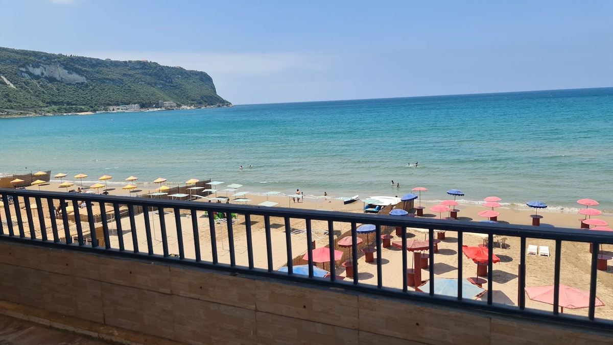 A scenic view of a beach is visible from the balcony, with soft waves lapping at the shore. Colorful umbrellas provide shade on the sandy beach, while distant figures enjoy the water. A lush green hill rises in the background under a clear blue sky.