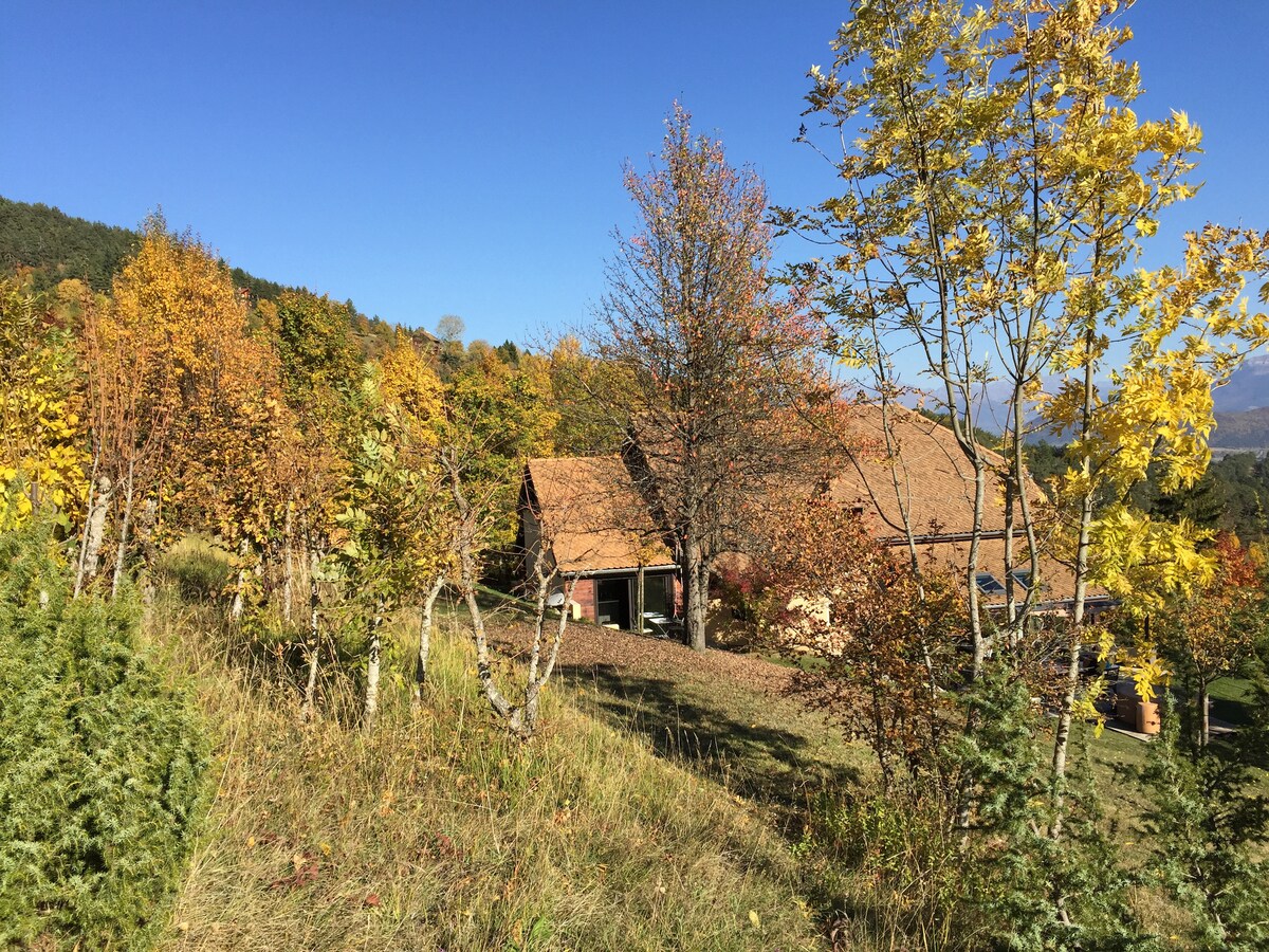 A serene landscape showcases a charming rustic building surrounded by trees displaying autumn foliage. The clear blue sky complements the warm colors of the leaves, while grassy slopes lead up to the structure, providing a peaceful atmosphere in the natural setting.