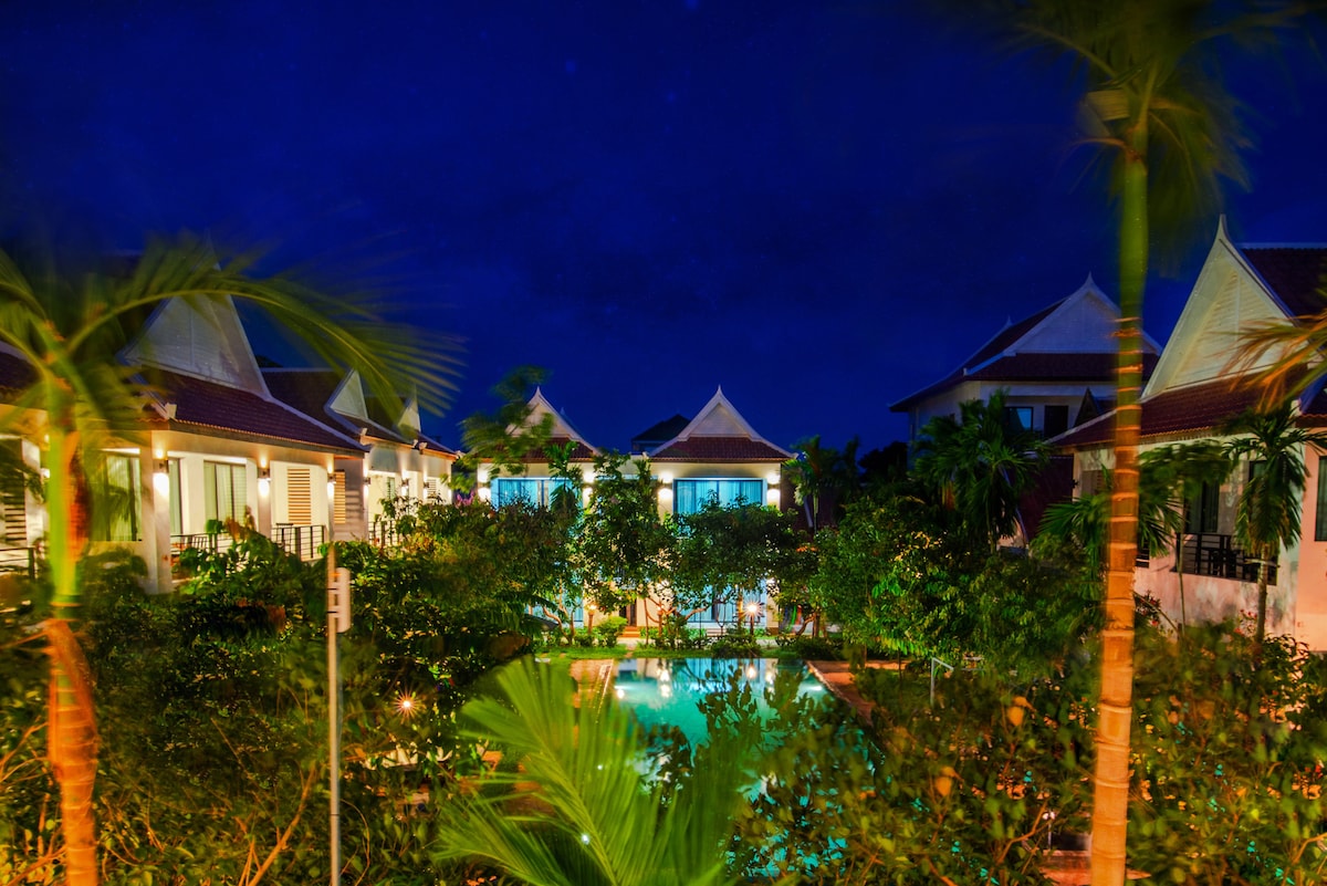 The view captures the serene ambiance of the resort at night, with well-lit villas surrounded by lush tropical vegetation. A pool lies at the center, reflecting the tranquil atmosphere, while palm trees gently sway in the breeze under the night sky.