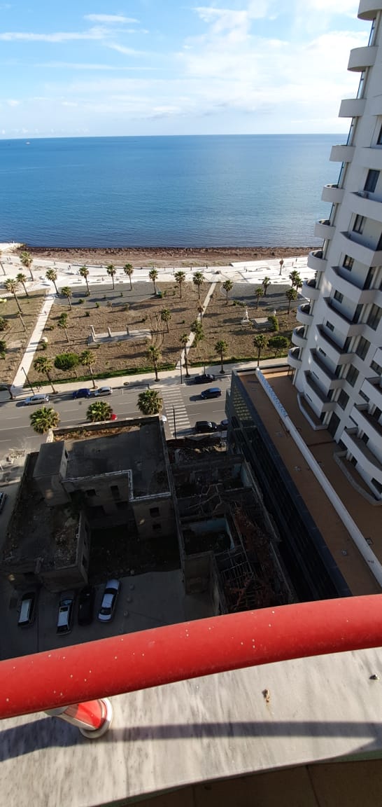 The view from the top floor showcases the coastline and beach area, with palm trees lining the walkway. A glimpse of the ocean is visible, along with the sandy shore. Below, a building structure and parked cars can be seen, situated along the street.