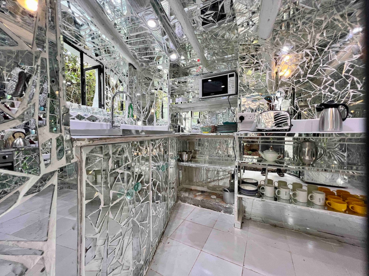 A uniquely designed kitchen showcases a mosaic of mirrored surfaces, reflecting light and color throughout the space. Modern appliances are visible, along with plates and cups neatly arranged on shelves. Natural light filters in through windows, enhancing the vibrant atmosphere.