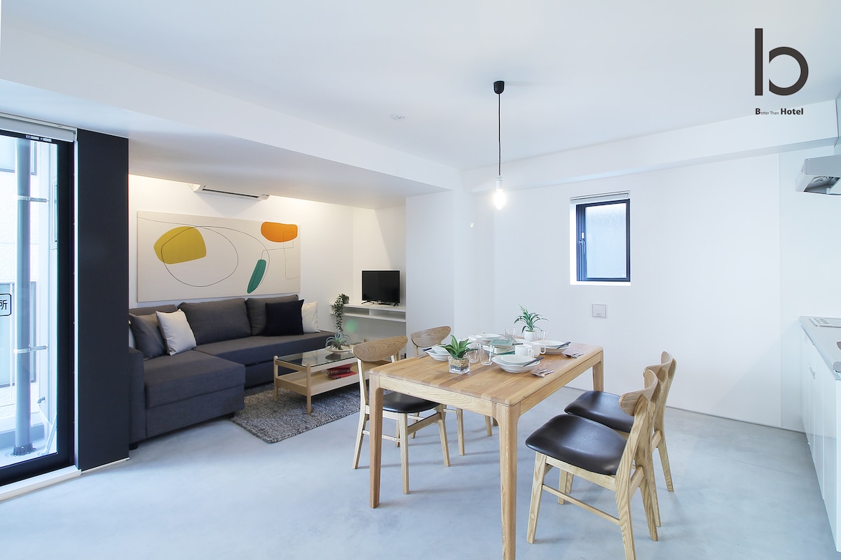 An open living area features a comfortable sofa, a dining table with four wooden chairs, and a wall-mounted TV. A window allows natural light to flow in, highlighting the minimalist decor and modern design.