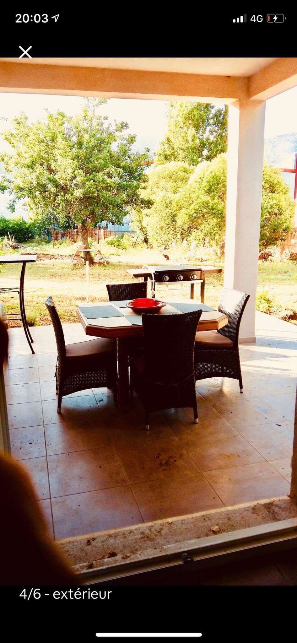 An outdoor dining area features a table set with four chairs, positioned on a tiled patio. A red centerpiece is placed on the table, while a barbecue grill is visible in the background. Lush greenery surrounds the area, creating a natural setting.