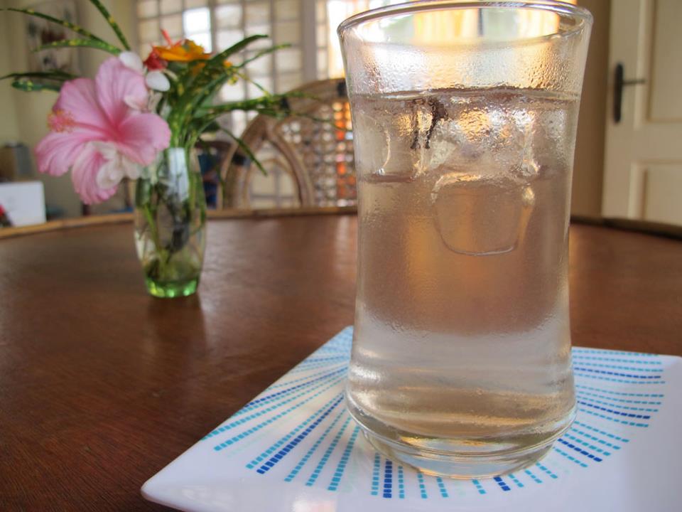 A glass of cold water with ice sits on a decorative plate, placed on a wooden table. In the background, a vase holds colorful flowers, and natural light filters through a window, creating a refreshing atmosphere.