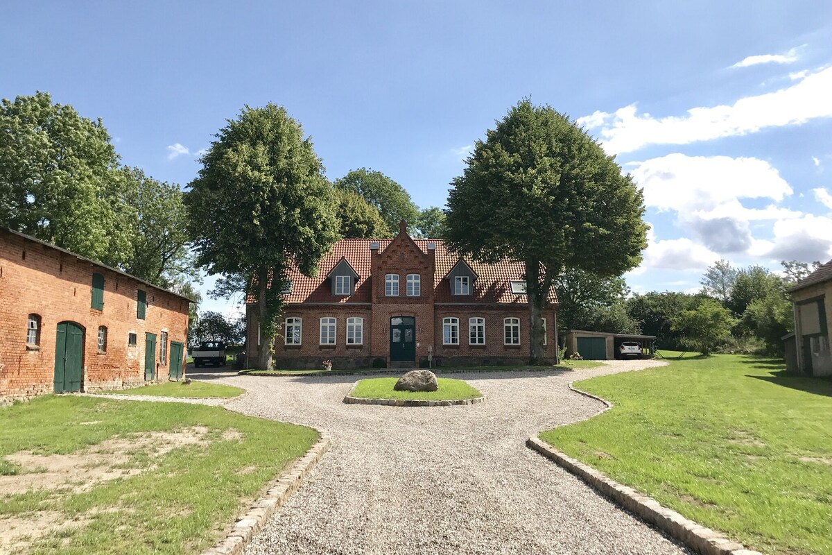 A traditional brick building is surrounded by well-maintained gardens and large trees. A gravel pathway leads to the entrance, showcasing a spacious layout. The sky is clear with a few clouds, suggesting a bright and sunny day.