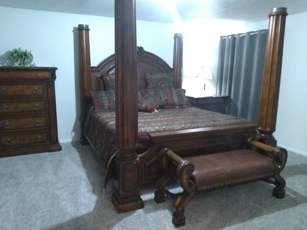 A spacious bedroom is highlighted by a king-sized four-poster bed with rich wood detailing. Decorative pillows complement the patterned bedding. A matching dresser stands nearby, while a bedside table holds a lamp. Soft carpeted flooring enhances comfort, and gray curtains frame a window.