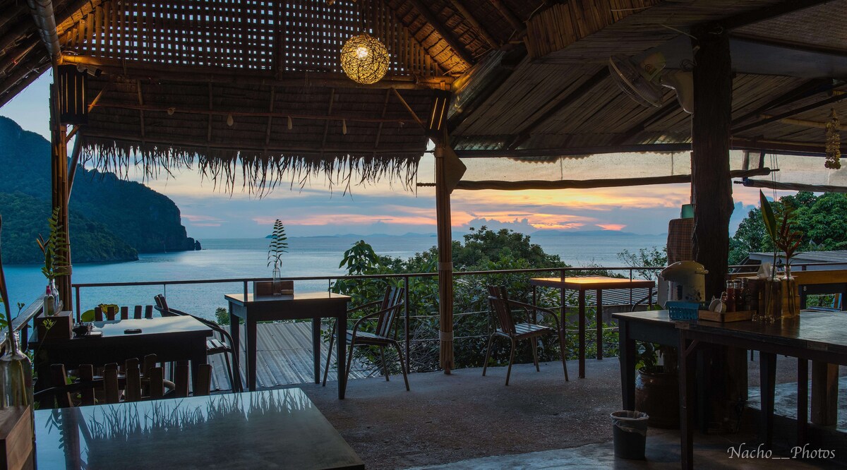 An open-air dining area is captured at dusk, featuring wooden tables and chairs arranged to overlook the sea. Natural materials like bamboo and thatch create a rustic charm, while vibrant colors of the sunset are visible in the sky, against a backdrop of lush hills and calm waters.