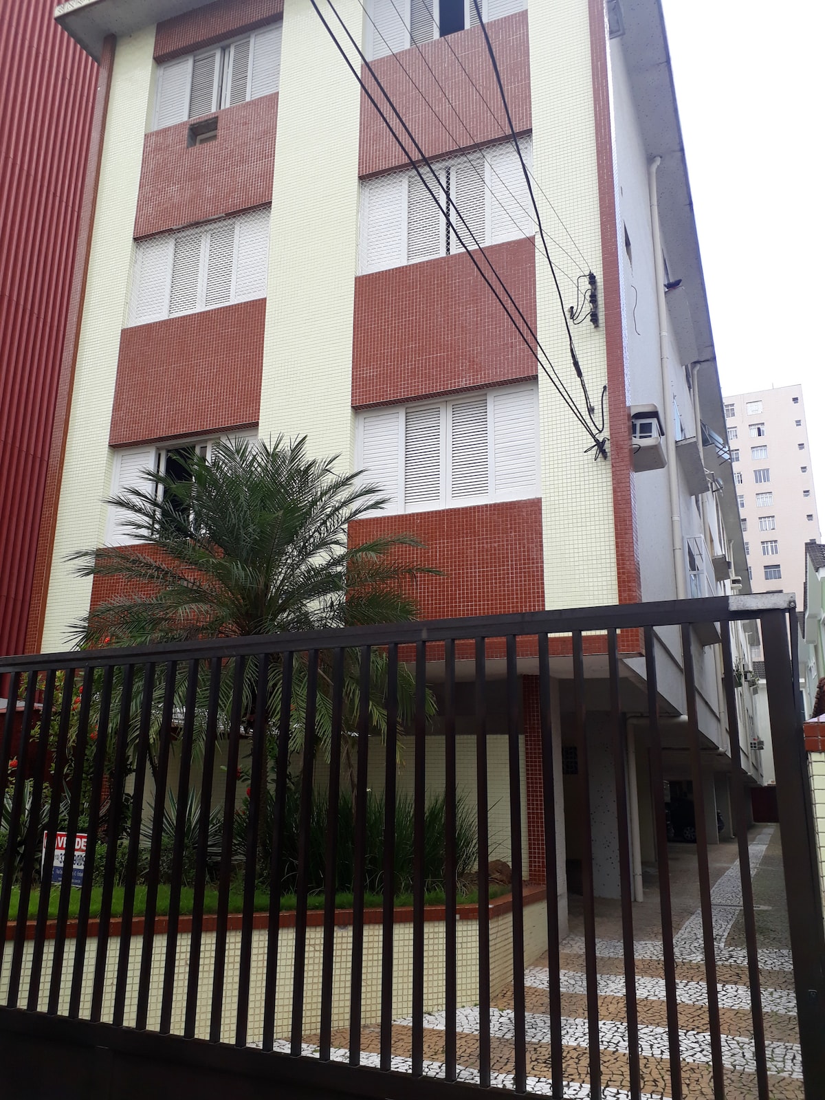 The apartment building features a modern design with a combination of red and cream exterior walls. Large windows allow natural light to enter, while a welcoming entrance is framed by greenery, including palms. A gated entry leads to the pathway lined with decorative tiles.