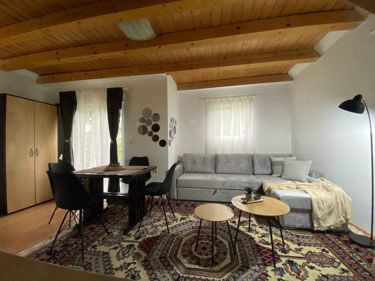 An open living area features a gray sectional sofa complemented by a light-colored throw and two circular coffee tables. A dining table is set for four, while decorative wall art circles add visual interest. Natural light filters through sheer curtains, enhancing the space.