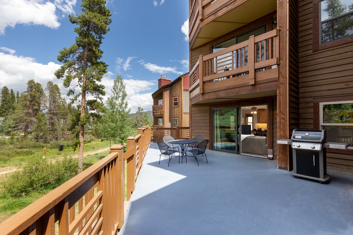 A large patio is shown, featuring a table with four chairs and a grill. Natural surroundings include trees and open space beyond the railing, alongside a condominium structure. A glass door leads into the living area, allowing for easy access to outdoor seating.