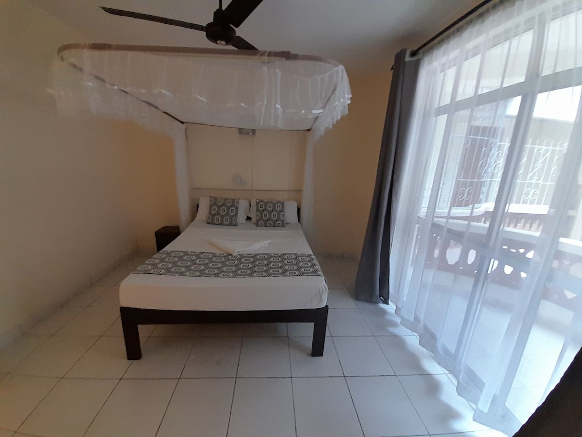 A spacious bed is framed by a decorative canopy and complemented by patterned pillows. Natural light filters through sheer curtains, illuminating the room, while a ceiling fan is positioned above. Sliding glass doors lead to a balcony area visible from the interior.