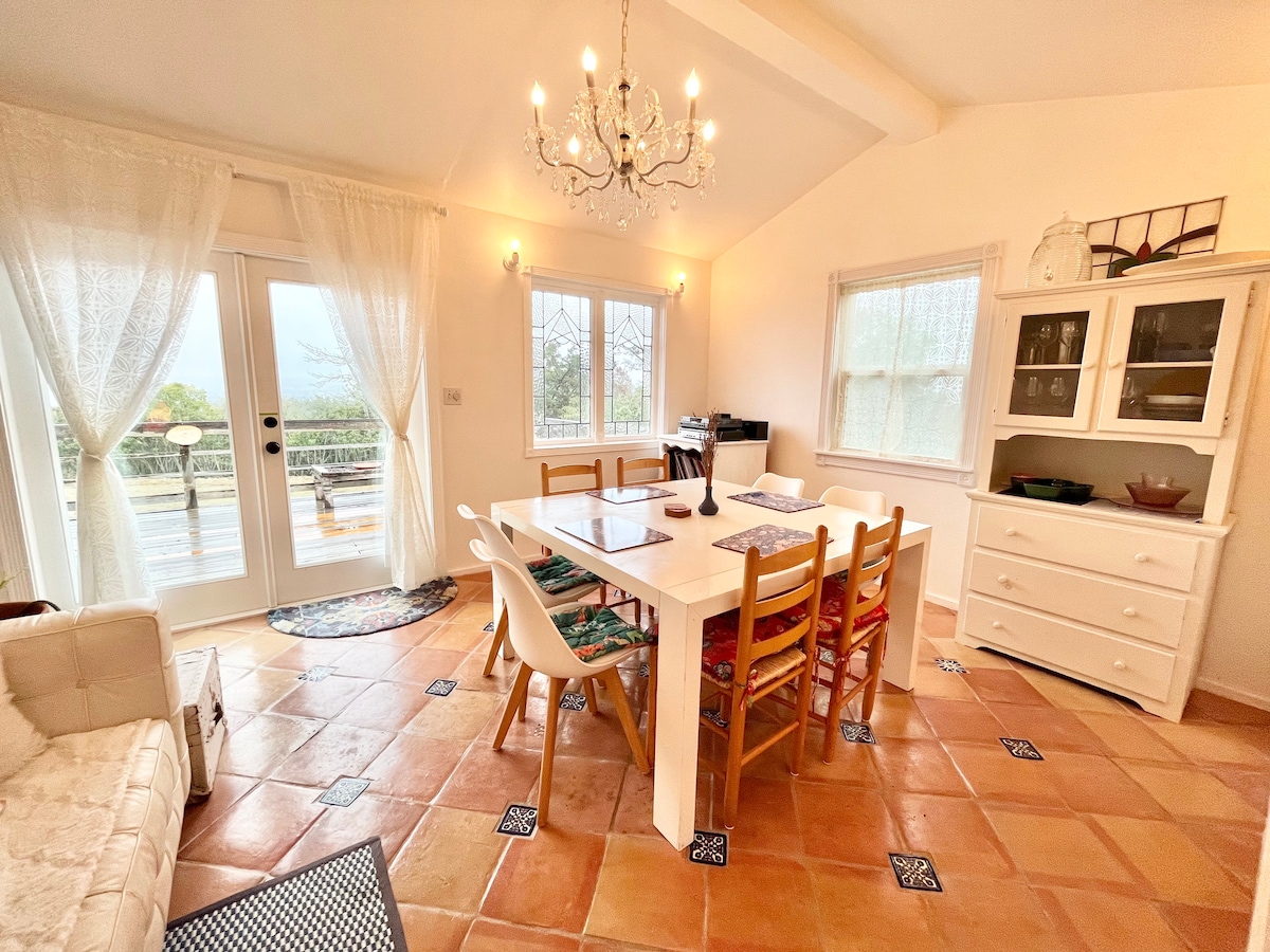 A bright dining area features a large white table surrounded by wooden chairs, each with colorful cushions. A chandelier hangs above, and sunlight streams through large windows with sheer curtains, illuminating the Saltillo tile floor. An inviting sideboard and a cozy seating area are included.