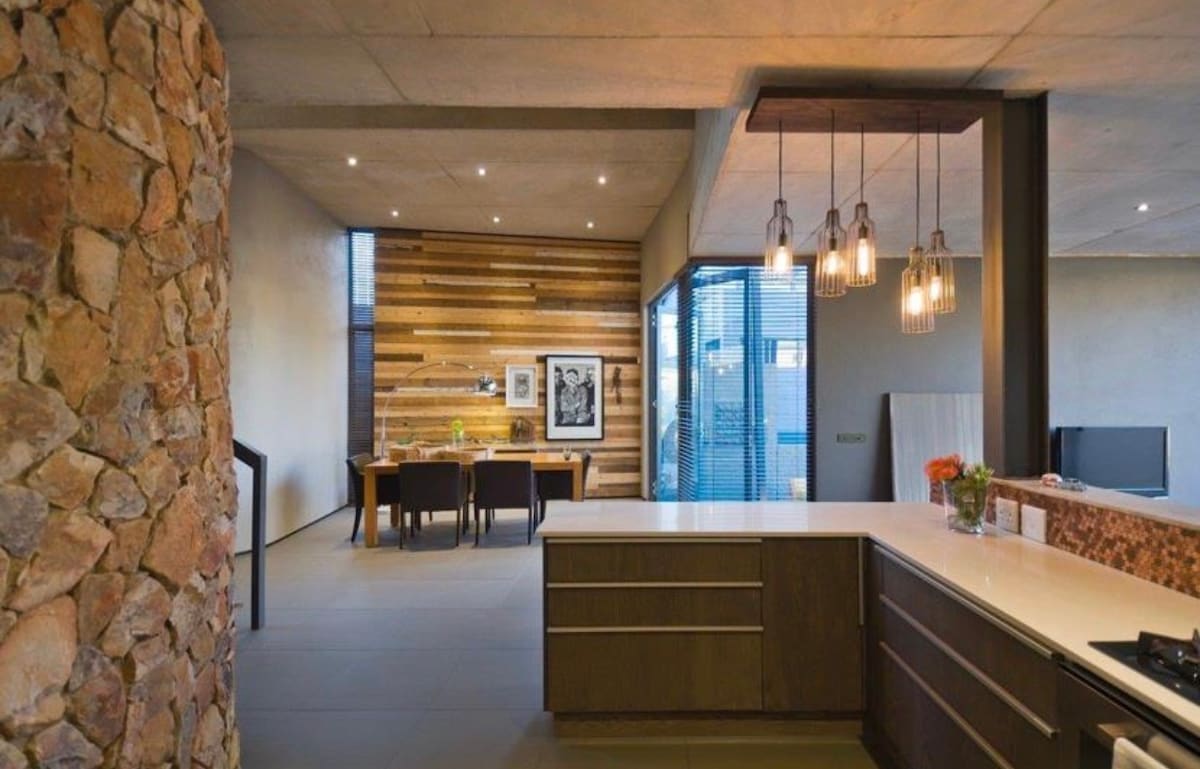 The open-plan kitchen features a spacious island with modern cabinetry and sleek countertops. A dining area with a wooden table and dark chairs is visible in the background, complemented by wood and stone accents on the walls. Natural light streams through large windows.