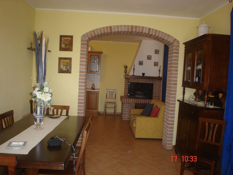 Living piano-terra con tavolo grande ed uno più piccolo, cucina e caminetto funzionante con divano.