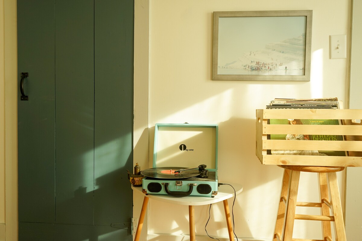 A vintage record player rests on a small white table, accompanied by a wooden crate filled with vinyl records. A blue-green door adds a pop of color, while natural light streams in, creating a warm ambiance in the corner of the room.