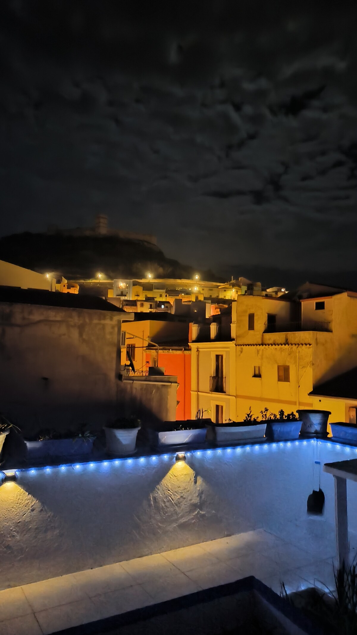 The nighttime view captures a charming town illuminated by warm lights. Distinct rooftops and buildings are visible, while a hill with a tower rises in the background. Decorative blue lights adorn the foreground, enhancing the inviting atmosphere of the scene.