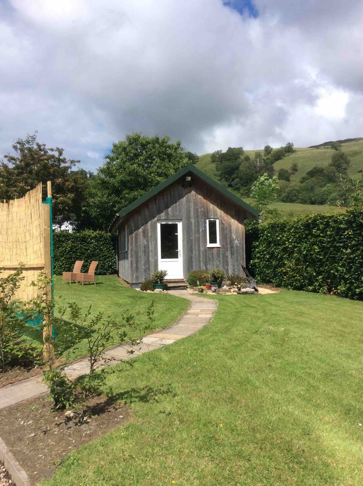 A quaint garden shed is situated among well-maintained grass and shrubs, showcasing a pathway leading to the entrance. Surrounding greenery provides privacy, while gentle hills are visible in the background beneath a partly cloudy sky.