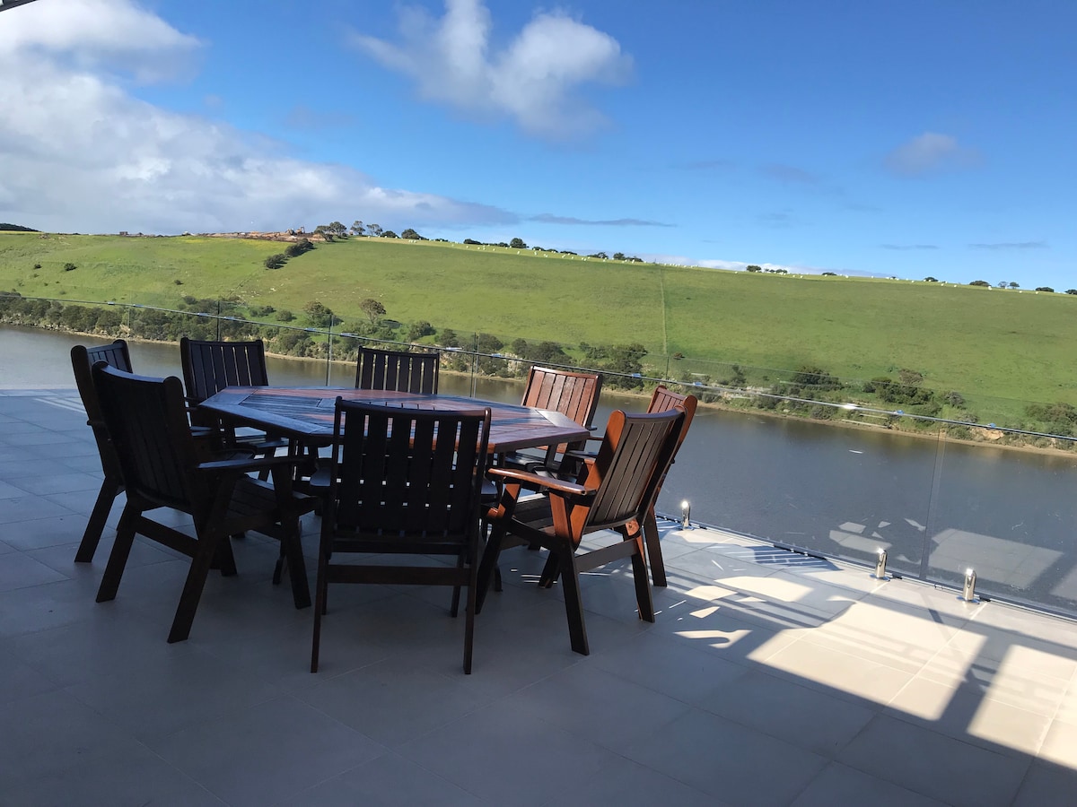 An outdoor dining area is presented with a large wooden table surrounded by eight matching chairs. The space offers views of a serene river against a backdrop of green hills, under a partly cloudy sky.