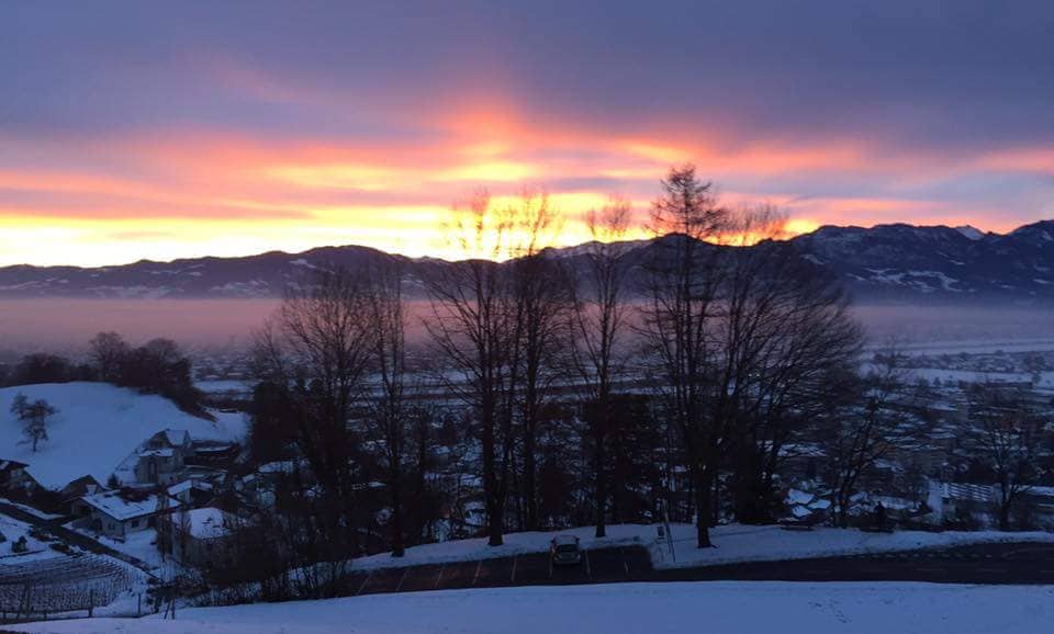 A serene winter landscape is displayed, featuring snow-covered ground and trees silhouetted against a vibrant sunset sky. The distant mountains can be seen in the background, while a peaceful valley lies beneath the soft mist, providing a tranquil view.