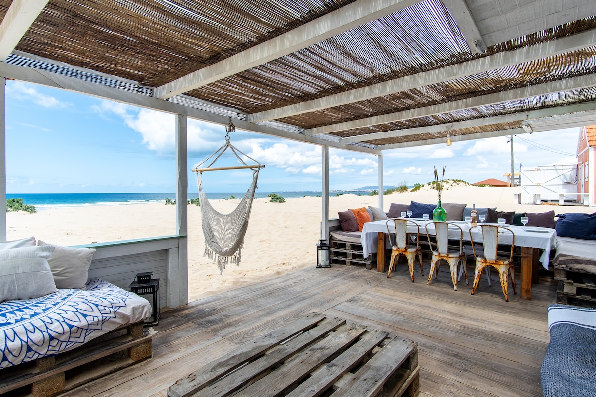 A shaded outdoor patio is presented with wooden flooring and overhead thatch. A hanging hammock is visible alongside a large dining table with wooden chairs. The space offers views of the sandy beach and ocean beyond, creating an inviting area for relaxation and gatherings.
