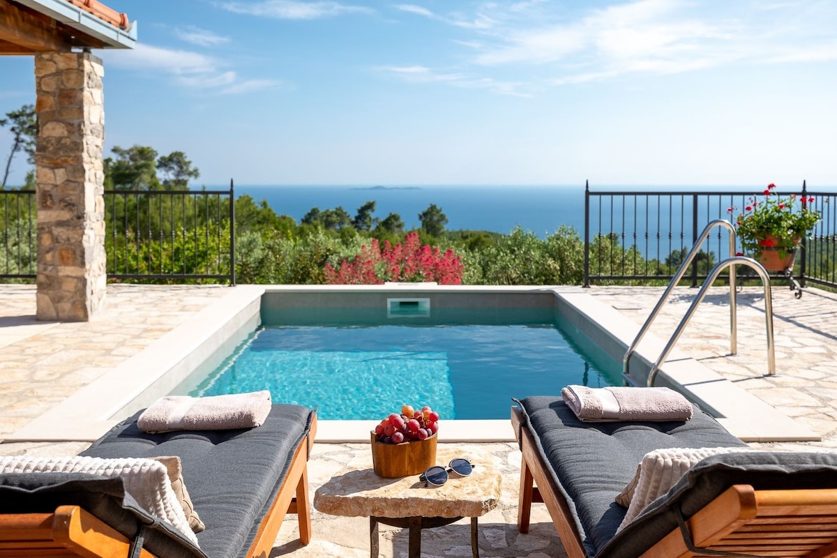 A small stone pool is situated on a sun-drenched terrace, surrounded by a stone patio. Two loungers face the tranquil water, accompanied by a small table holding a bowl of fruit and sunglasses. Lush greenery and the sea are visible in the background.
